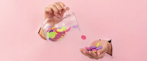 fundo-rosa-duas-maos-brancas-uma-segura-jarra-moedas-coloridas-outra-segura-moedas-coloridas-header